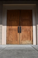A pair of large wooden double doors with vertical panels and two dark metal handles. The doors are set in a light-colored frame, and the surrounding area includes textured brick walls and a concrete floor.