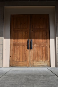 A pair of large wooden double doors with vertical panels and two dark metal handles. The doors are set in a light-colored frame, and the surrounding area includes textured brick walls and a concrete floor.