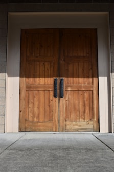 A pair of large wooden double doors with vertical panels and two dark metal handles. The doors are set in a light-colored frame, and the surrounding area includes textured brick walls and a concrete floor.