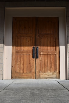 A pair of large wooden double doors with vertical panels and two dark metal handles. The doors are set in a light-colored frame, and the surrounding area includes textured brick walls and a concrete floor.