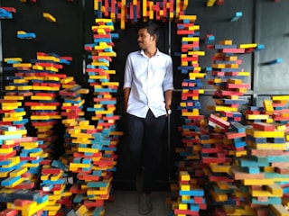 A man in a white shirt and black pants stands amidst a creative installation of colorful, stacked blocks. The blocks are predominantly red, yellow, and blue, arranged chaotically around him forming a passage.