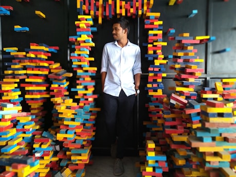 A man in a white shirt and black pants stands amidst a creative installation of colorful, stacked blocks. The blocks are predominantly red, yellow, and blue, arranged chaotically around him forming a passage.