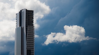 A tall, modern skyscraper with a sleek design stands prominently against a dramatic sky filled with thick, fluffy white clouds. The building features a blend of glass and concrete, and its vertical lines contrast sharply with the softness of the clouds.