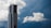 A tall, modern skyscraper with a sleek design stands prominently against a dramatic sky filled with thick, fluffy white clouds. The building features a blend of glass and concrete, and its vertical lines contrast sharply with the softness of the clouds.