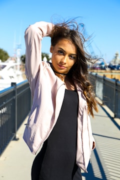 A woman with long, flowing hair poses confidently outdoors on a sunny day. She is wearing a light pink jacket over a black dress, and stands on a walkway lined with a black metal railing. Her hand is lifting her hair, and she gazes directly at the camera, with boats and a clear blue sky in the background.