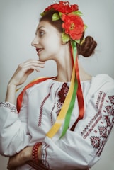 A person is dressed in traditional attire, featuring a white embroidered blouse with red patterns and a headdress adorned with bright red flowers and multicolored ribbons. The hair is styled in a bun, and the person has a contemplative expression.