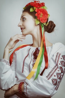 A person is dressed in traditional attire, featuring a white embroidered blouse with red patterns and a headdress adorned with bright red flowers and multicolored ribbons. The hair is styled in a bun, and the person has a contemplative expression.
