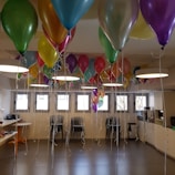 A brightly lit room filled with an abundance of colorful balloons tied with white strings. The balloons are suspended from the ceiling, creating a festive atmosphere. The room contains several black chairs and tables with items on them. Large rectangular windows allow natural light to illuminate the space.