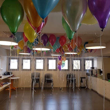 A brightly lit room filled with an abundance of colorful balloons tied with white strings. The balloons are suspended from the ceiling, creating a festive atmosphere. The room contains several black chairs and tables with items on them. Large rectangular windows allow natural light to illuminate the space.