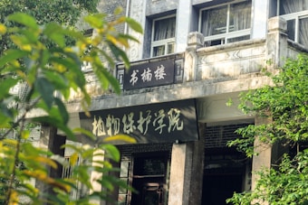 The image features the entrance of a concrete building with a prominent sign displaying Chinese characters. The structure has a slightly aged appearance with visible windows and a balcony. Greenery surrounds the area, with branches and leaves partially obscuring the view of the building.