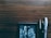 A wooden surface with a small framed black and white painting in the lower left corner and three pens of different styles laid next to it. The pens include a silver one, a black one with gold accents, and a clear plastic one. The painting depicts various geometric shapes and reflections.