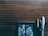 A wooden surface with a small framed black and white painting in the lower left corner and three pens of different styles laid next to it. The pens include a silver one, a black one with gold accents, and a clear plastic one. The painting depicts various geometric shapes and reflections.