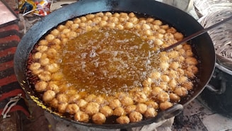 A large black pan filled with oil is being used to fry numerous small, round dough pieces. The dough pieces are golden brown and are evenly distributed around the pan's edge. Bubbles can be seen rising in the oil, indicating active frying. A ladle or skimmer is partially visible, possibly used to stir or remove the fried items. Nearby, there are baskets that may be used for holding or serving the fried items.