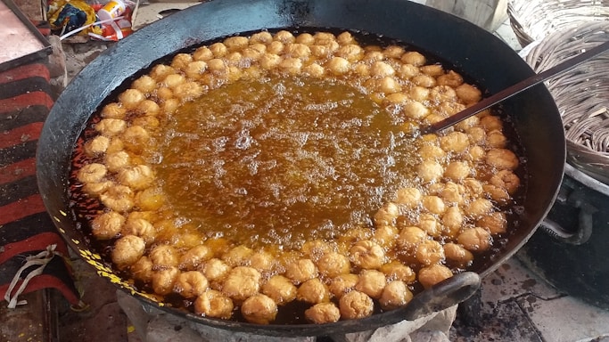 A large black pan filled with oil is being used to fry numerous small, round dough pieces. The dough pieces are golden brown and are evenly distributed around the pan's edge. Bubbles can be seen rising in the oil, indicating active frying. A ladle or skimmer is partially visible, possibly used to stir or remove the fried items. Nearby, there are baskets that may be used for holding or serving the fried items.