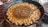 A large black pan filled with oil is being used to fry numerous small, round dough pieces. The dough pieces are golden brown and are evenly distributed around the pan's edge. Bubbles can be seen rising in the oil, indicating active frying. A ladle or skimmer is partially visible, possibly used to stir or remove the fried items. Nearby, there are baskets that may be used for holding or serving the fried items.