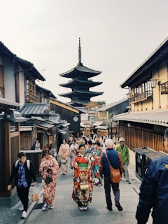 A street scene in a traditional Japanese setting with numerous people walking. Many individuals are wearing colorful kimonos, and traditional wooden buildings line both sides of the path. A prominent pagoda is visible in the background, enhancing the cultural atmosphere.