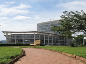 A modern multi-story building with a glass facade is surrounded by a lush green landscape. In the foreground, a brick path leads to a yellow bench placed on well-manicured grass. The sky above is partly cloudy, and the overall scene is peaceful and orderly.