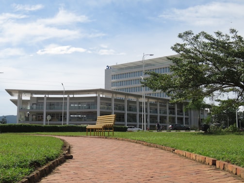 A modern multi-story building with a glass facade is surrounded by a lush green landscape. In the foreground, a brick path leads to a yellow bench placed on well-manicured grass. The sky above is partly cloudy, and the overall scene is peaceful and orderly.
