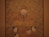 A traditional Asian painting features a central figure with a long beard and ornate headdress holding a banner. The figure is surrounded by smaller faces and intricate background patterns resembling clouds. The colors are predominantly earthy tones with a slightly faded appearance, suggesting age.