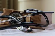 A stethoscope rests on a white surface alongside a sphygmomanometer. Behind them, a thick book with a blue cover and a brown fabric bag are visible. The focus is on the medical instruments, suggesting a clinical or healthcare context.