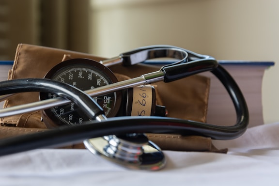 A stethoscope rests on a white surface alongside a sphygmomanometer. Behind them, a thick book with a blue cover and a brown fabric bag are visible. The focus is on the medical instruments, suggesting a clinical or healthcare context.