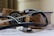 A stethoscope rests on a white surface alongside a sphygmomanometer. Behind them, a thick book with a blue cover and a brown fabric bag are visible. The focus is on the medical instruments, suggesting a clinical or healthcare context.