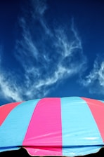 A colorful striped canopy or umbrella occupies the lower part of the image, with alternating pink and blue stripes. Above it, a backdrop of deep blue sky features wispy, partially translucent clouds, creating a striking contrast between the canopy and the sky.