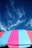 A colorful striped canopy or umbrella occupies the lower part of the image, with alternating pink and blue stripes. Above it, a backdrop of deep blue sky features wispy, partially translucent clouds, creating a striking contrast between the canopy and the sky.