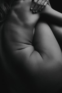 A black and white photograph featuring a nude body, focusing on the skin's texture and contours. The composition captures the natural curves and shadows, with a hand gently placed on the body, contributing to the intimate and artistic quality of the image.