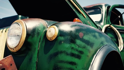 A close-up view of an aged, vintage car, primarily dark green with patches of rust and wear. The focus is on the rounded front fender and the two headlights, one of which is a small amber indicator light. The car's hood is open, suggesting possible maintenance or inspection. The paint displays a patina effect indicative of long-term exposure to the elements.