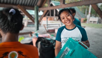 A person is smiling and holding a translucent blue plastic container. They are wearing a T-shirt with text that reads 'College of Fisheries.' The setting appears to be a covered outdoor area with wooden beams and picnic tables in the background. Another person is partially visible, wearing an orange T-shirt, with their back to the camera.