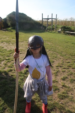 A child dressed in playful warrior attire stands on a grassy field. The child is wearing a helmet with a face guard and is holding a long staff or spear. Their outfit includes a shirt with a lemon graphic, a tied gray polka-dot sweater around the waist, patterned leggings, and red boots. The background features a structure made of wooden beams and bushes under a clear blue sky.