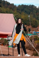 A person is standing outdoors with a backdrop of trees and what appears to be a tent. They are dressed in a black outfit with an orange jacket tied around their waist and a white scarf draped over their shoulders. The setting suggests an outdoor camping or nature scene.