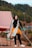 A person is standing outdoors with a backdrop of trees and what appears to be a tent. They are dressed in a black outfit with an orange jacket tied around their waist and a white scarf draped over their shoulders. The setting suggests an outdoor camping or nature scene.
