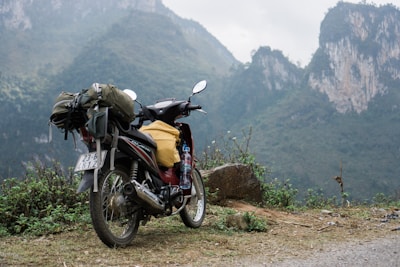 A motorcycle loaded with travel gear including a backpack and a plastic bottle stands on a dirt path. The background features rugged, misty mountains covered with green foliage.