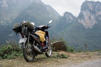 A motorcycle loaded with travel gear including a backpack and a plastic bottle stands on a dirt path. The background features rugged, misty mountains covered with green foliage.