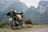 A motorcycle loaded with travel gear including a backpack and a plastic bottle stands on a dirt path. The background features rugged, misty mountains covered with green foliage.