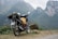 A motorcycle loaded with travel gear including a backpack and a plastic bottle stands on a dirt path. The background features rugged, misty mountains covered with green foliage.
