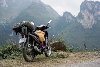 A motorcycle loaded with travel gear including a backpack and a plastic bottle stands on a dirt path. The background features rugged, misty mountains covered with green foliage.