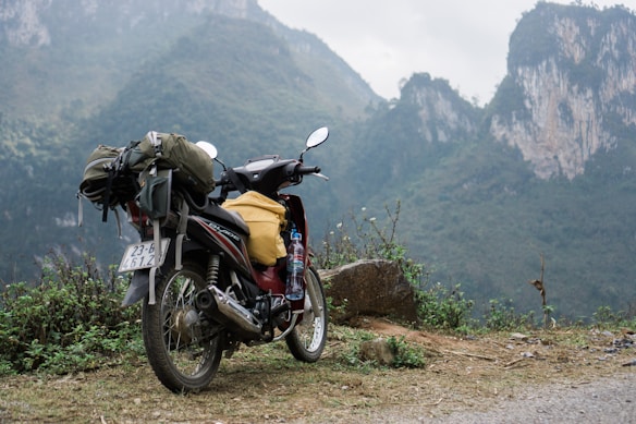 A motorcycle loaded with travel gear including a backpack and a plastic bottle stands on a dirt path. The background features rugged, misty mountains covered with green foliage.