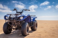 A blue all-terrain vehicle (ATV) is parked on an expansive sandy desert. The vehicle has large rugged tires and a sturdy frame, with handlebars equipped with control levers. The sky above is clear with some light clouds, creating a bright and open atmosphere.