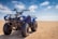 A blue all-terrain vehicle (ATV) is parked on an expansive sandy desert. The vehicle has large rugged tires and a sturdy frame, with handlebars equipped with control levers. The sky above is clear with some light clouds, creating a bright and open atmosphere.
