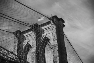 An architectural view of a large suspension bridge, focusing on the stone tower and the intricate web of cables. A U.S. flag is mounted on top of the tower, fluttering in the breeze. The image is captured in black and white, highlighting the textures of the stone and metal structures.
