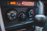 The image captures the control panel of a car interior, featuring air conditioning and heating dials. The panel includes an array of adjustable knobs with symbols indicating various settings like airflow direction and temperature. The digital clock reads 6:22, and there is a triangular hazard light button positioned above the controls.