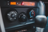 The image captures the control panel of a car interior, featuring air conditioning and heating dials. The panel includes an array of adjustable knobs with symbols indicating various settings like airflow direction and temperature. The digital clock reads 6:22, and there is a triangular hazard light button positioned above the controls.