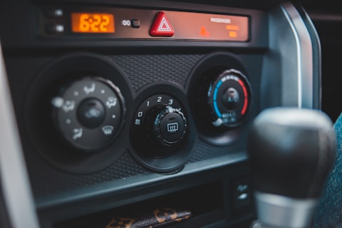 The image captures the control panel of a car interior, featuring air conditioning and heating dials. The panel includes an array of adjustable knobs with symbols indicating various settings like airflow direction and temperature. The digital clock reads 6:22, and there is a triangular hazard light button positioned above the controls.