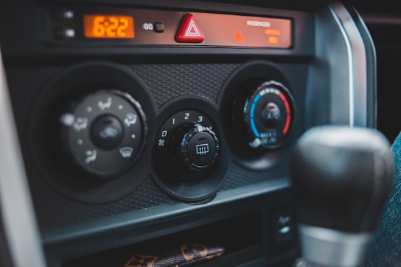 The image captures the control panel of a car interior, featuring air conditioning and heating dials. The panel includes an array of adjustable knobs with symbols indicating various settings like airflow direction and temperature. The digital clock reads 6:22, and there is a triangular hazard light button positioned above the controls.