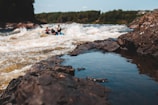 A fast-flowing river with white water rapids navigated by people in a raft. The foreground features rocky riverbanks with water reflections. Lush greenery and dense trees line the background.