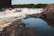 A fast-flowing river with white water rapids navigated by people in a raft. The foreground features rocky riverbanks with water reflections. Lush greenery and dense trees line the background.