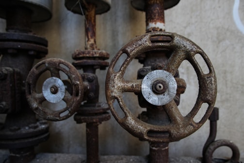 Rusty metal valves with circular handles are positioned against a textured wall. The valves show signs of wear and aging, with visible rust and corroded surfaces.